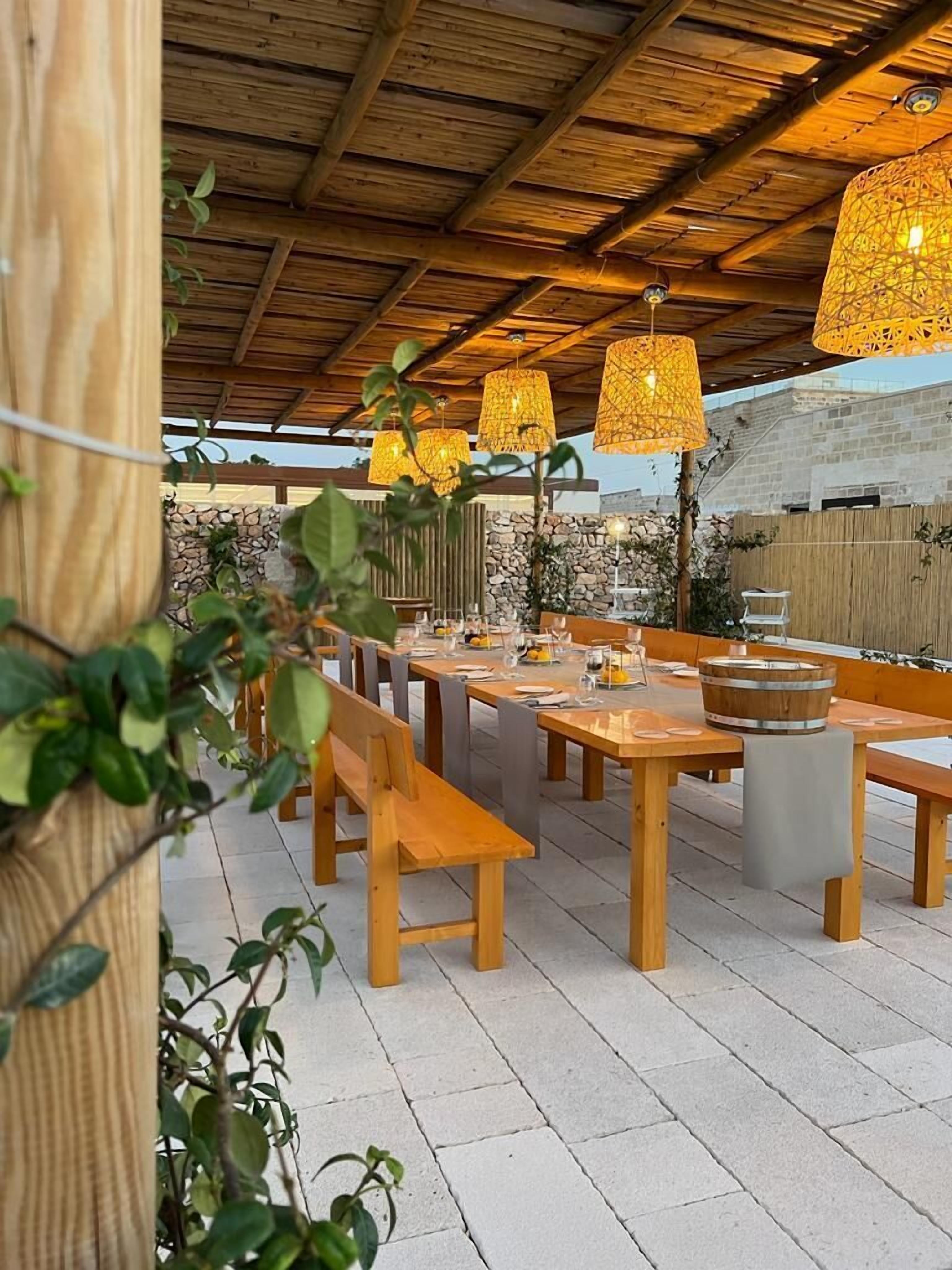 Un patio esterno con tavoli e panche in legno, circondato da muri in pietra e vegetazione.