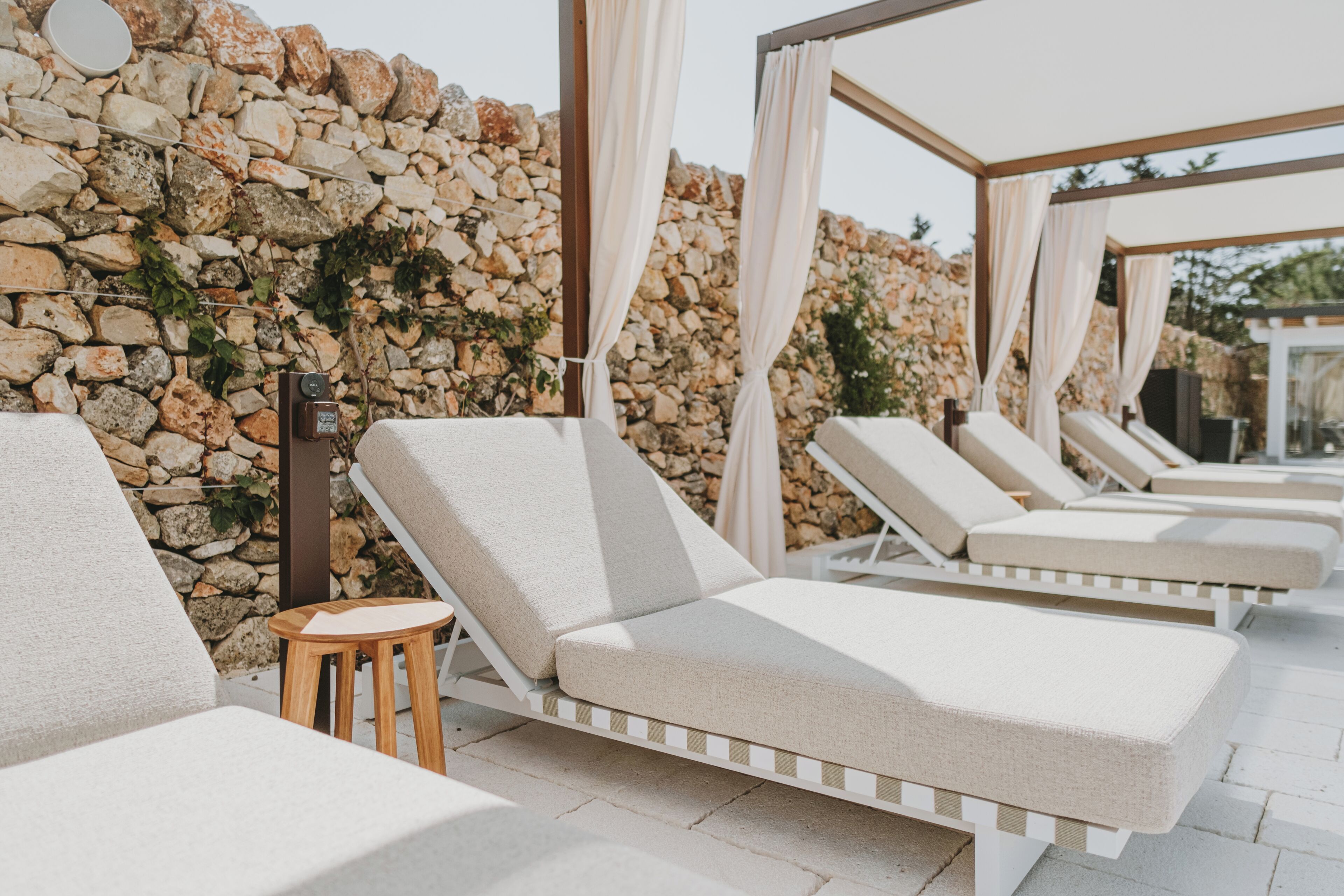 A sunlit outdoor lounge area with stone walls and cushioned lounge chairs.