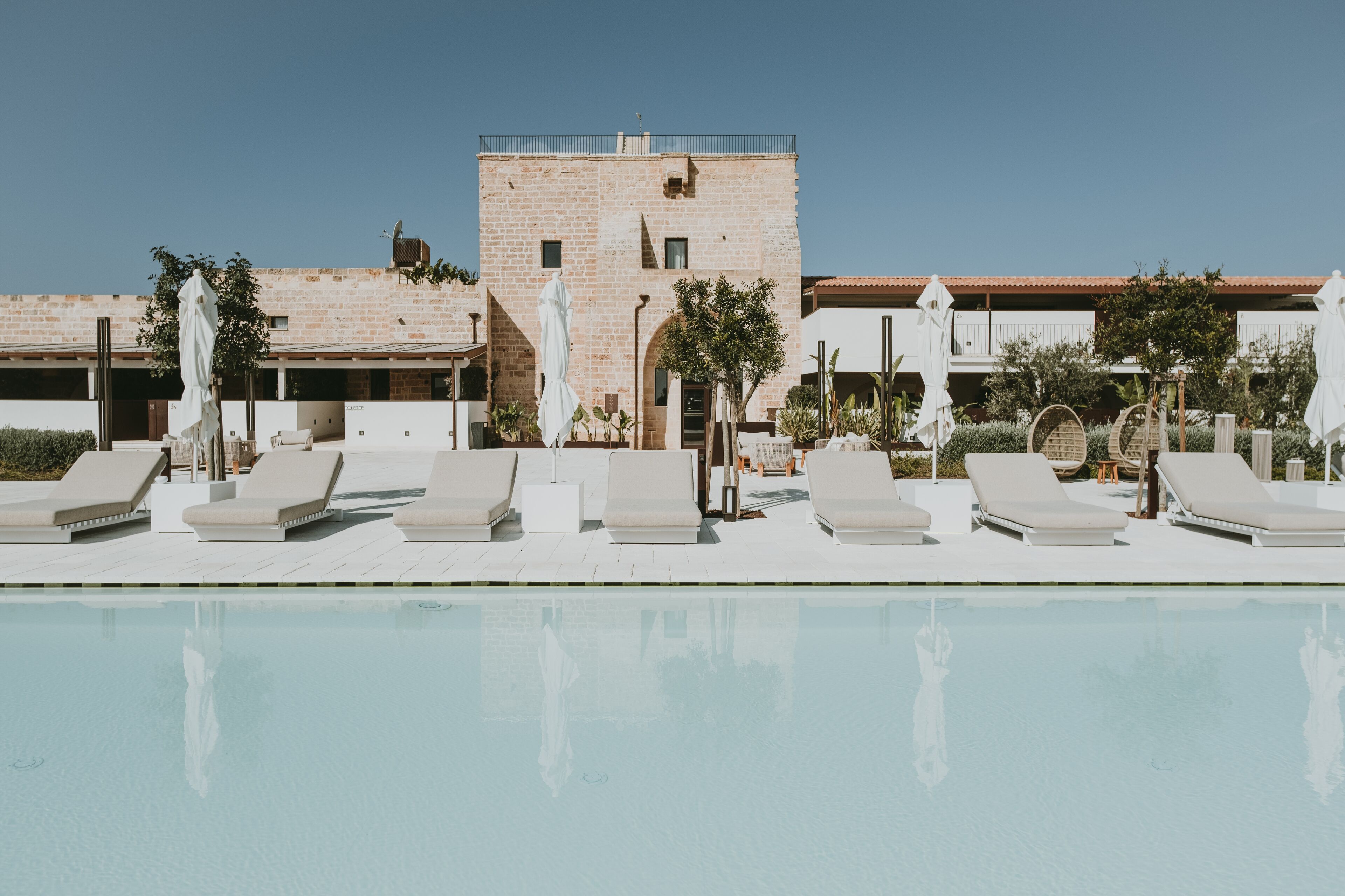 A poolside area with lounge chairs and umbrellas, a brick building, and a clear sky.