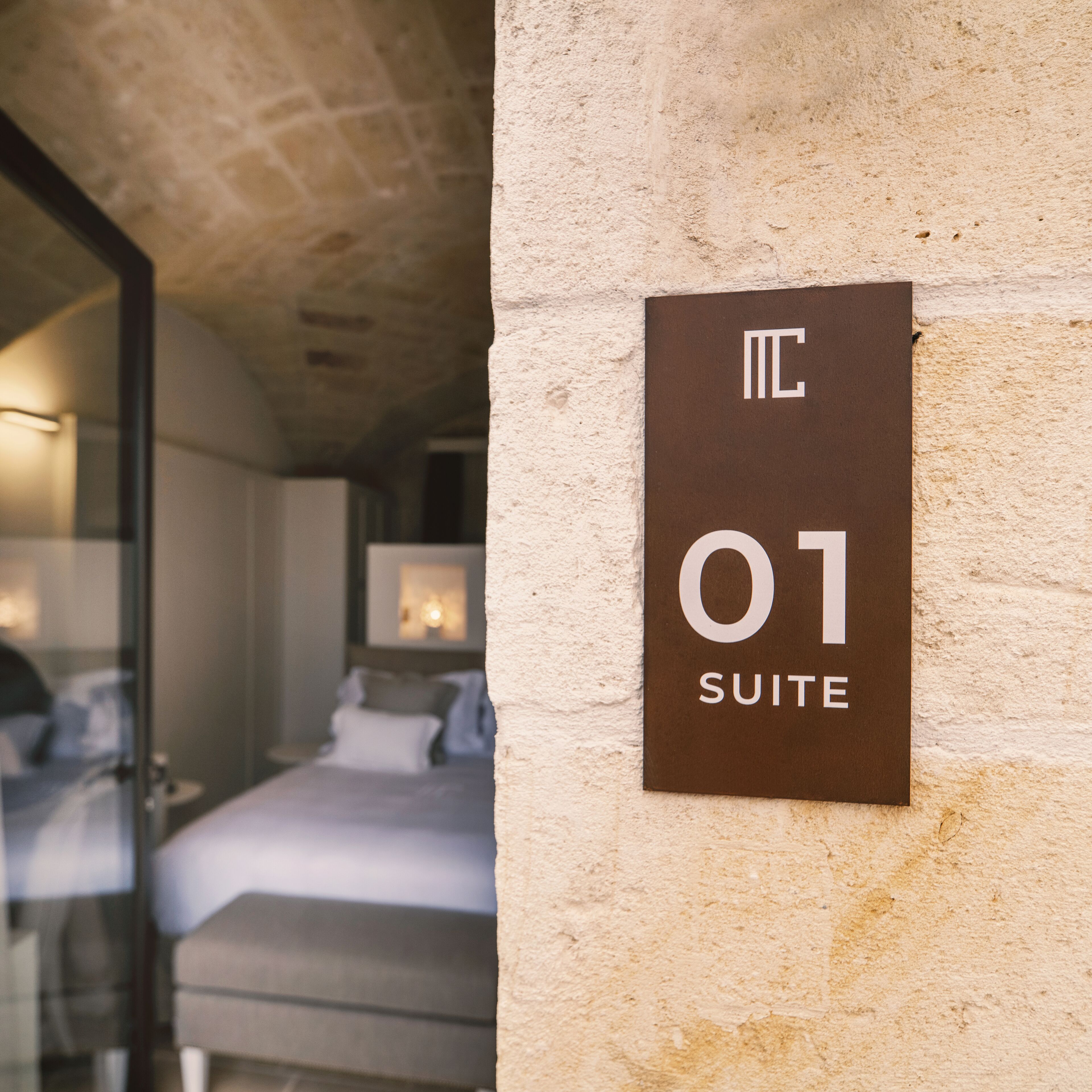 Letrero de hotel que indica la habitación número 01, suite, con una cama visible y una puerta de vidrio que da a otra habitación.