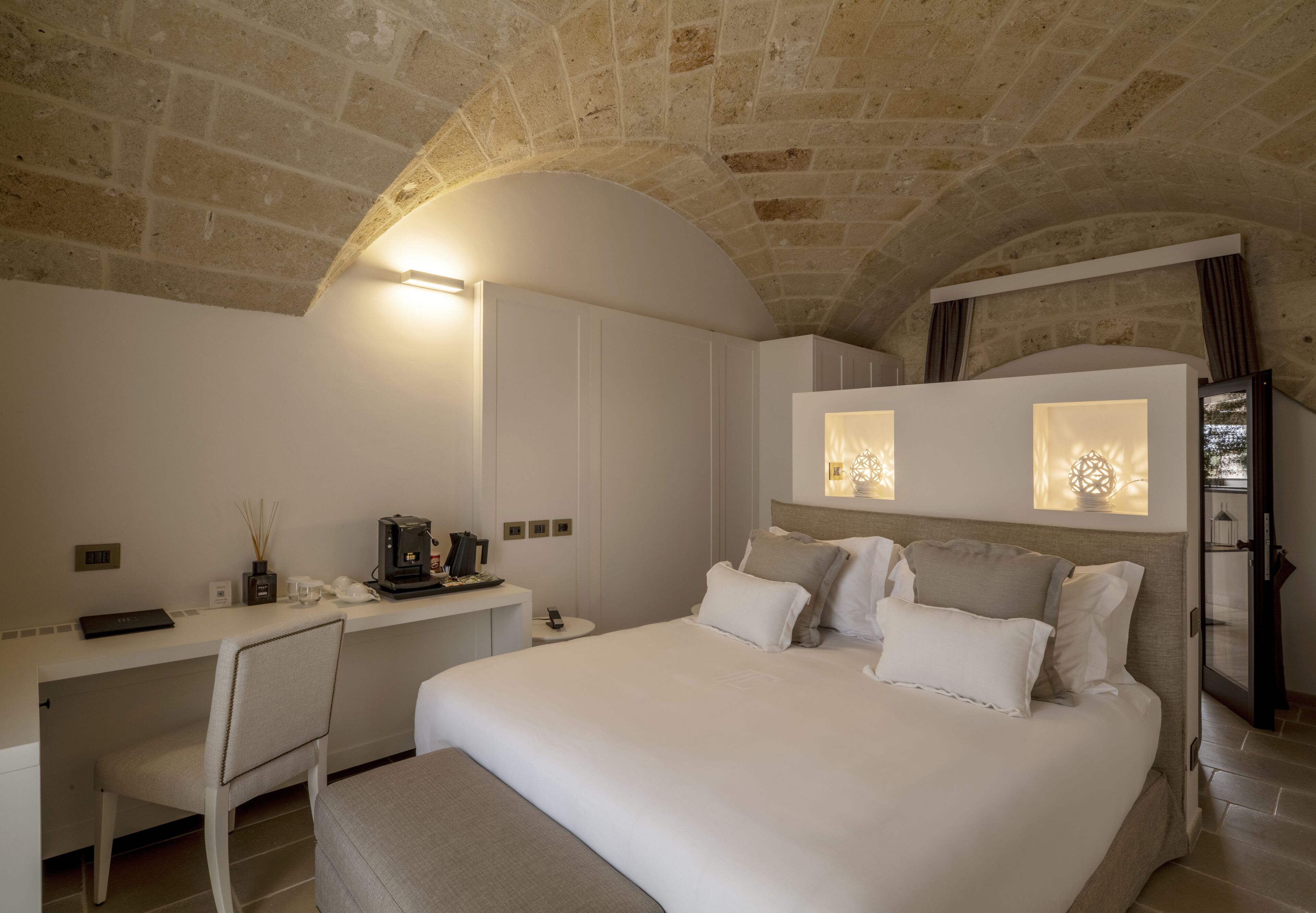 Een moderne hotelkamer met een stenen plafond, een groot bed met veel kussens, een bureau met stoel en een bijzettafeltje met een telefoon en een lamp.