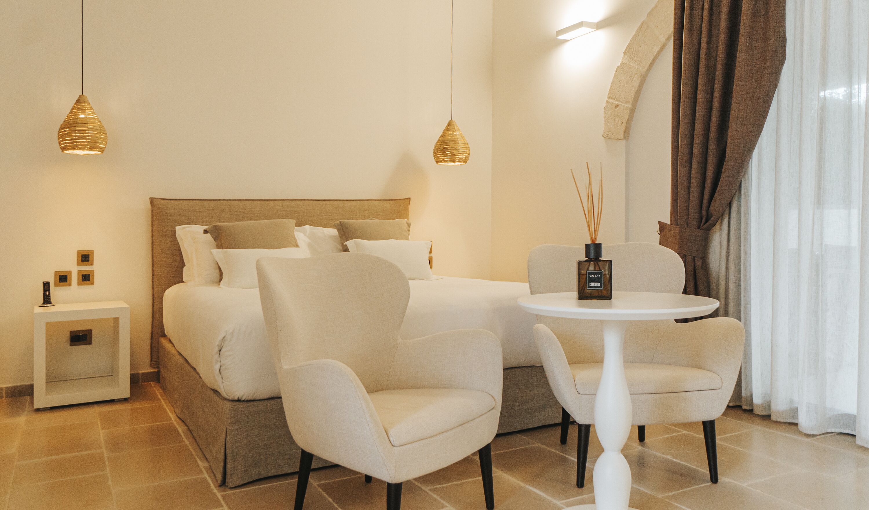 A hotel room with a bed, two chairs, a small table, and two pendant lights.