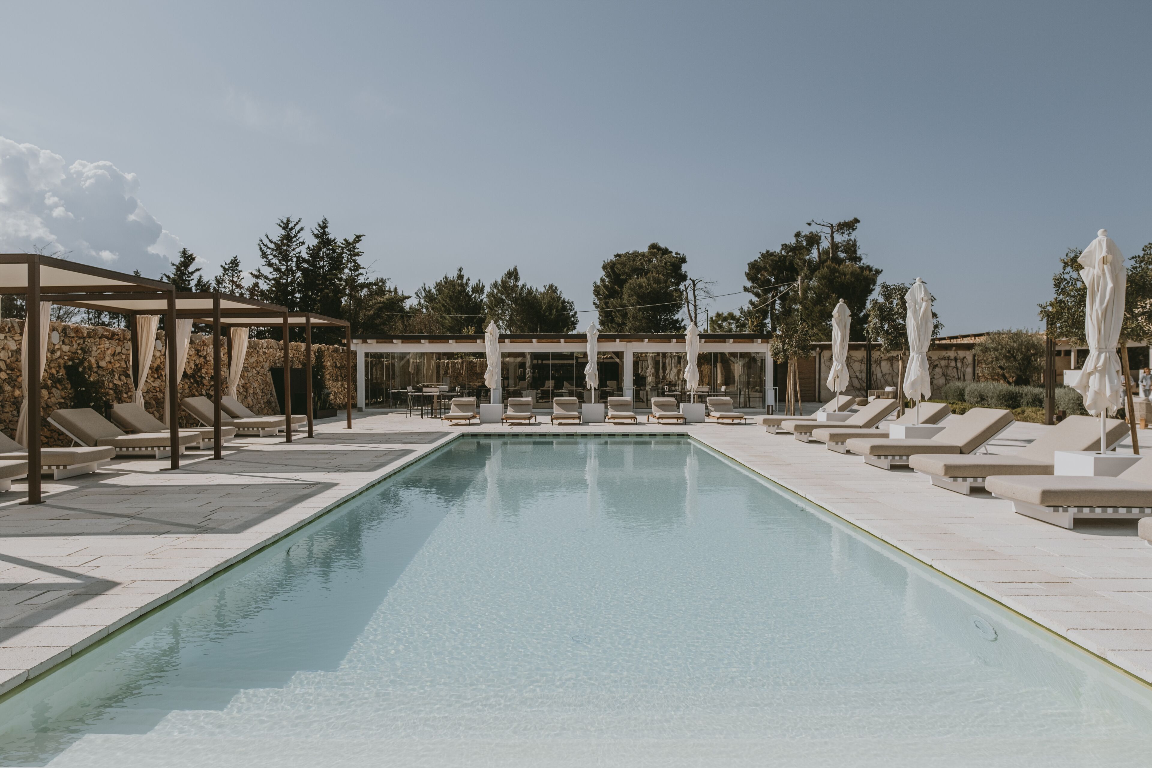 Una piscina rectangular con sillas de descanso y sombrillas, rodeada por un muro de piedra y árboles.