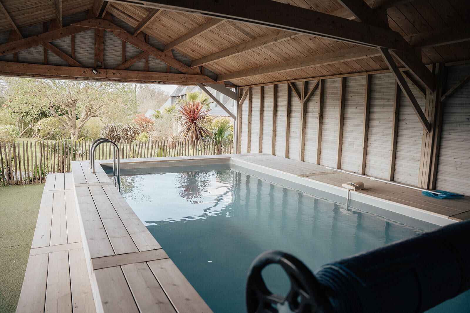 An indoor swimming pool with a wooden deck, a diving board, and a covered roof structure.
