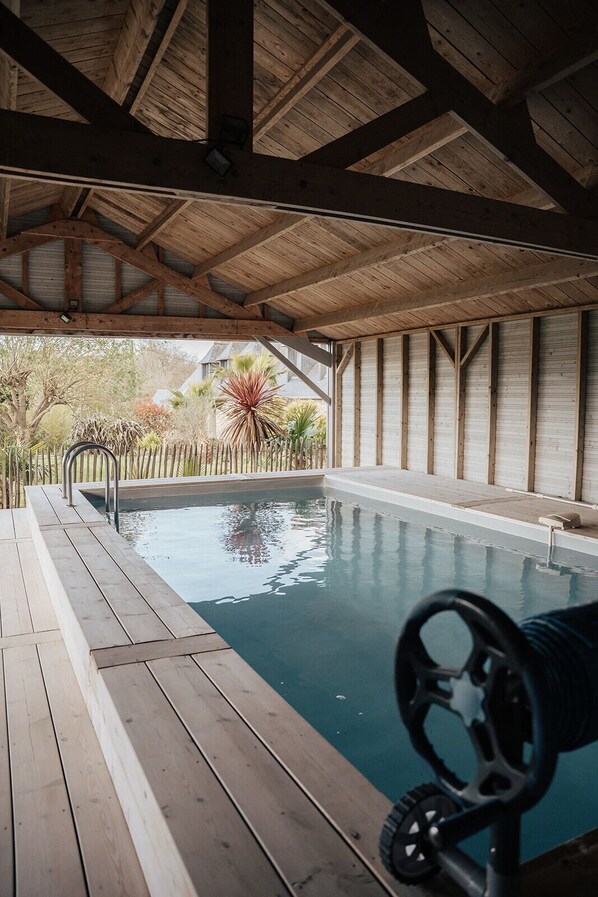 An indoor swimming pool with a wooden deck, metal handrail, and a view of a garden with various plants.