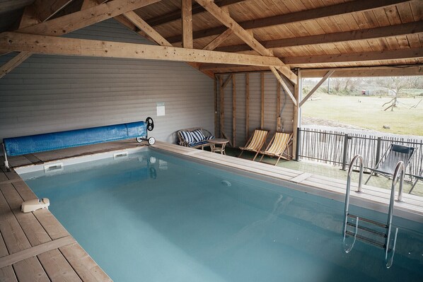 Una piscina interior con vigas de madera, sillas de descanso y vistas al exterior.