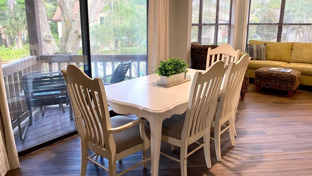 Un comedor con una mesa y sillas blancas, una planta pequeña sobre la mesa, y vistas al exterior a través de grandes ventanales.