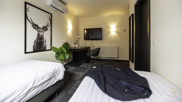 A hotel room with two beds, a desk, a chair, a TV, and a radiator. - Smart Bnb - Hotel Battice (Herve)