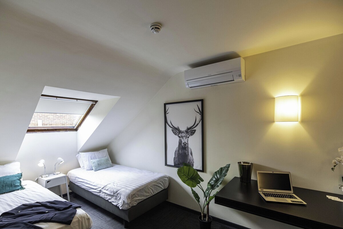 Una habitación de hotel moderna con una cama, un escritorio con una computadora portátil, una planta en maceta y un aire acondicionado instalado en la pared.