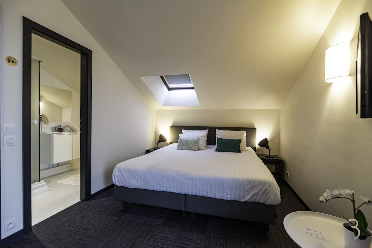 Una habitación de hotel moderna con tragaluz, una cama grande, lámparas de noche, un baño con lavabo y un espejo.
