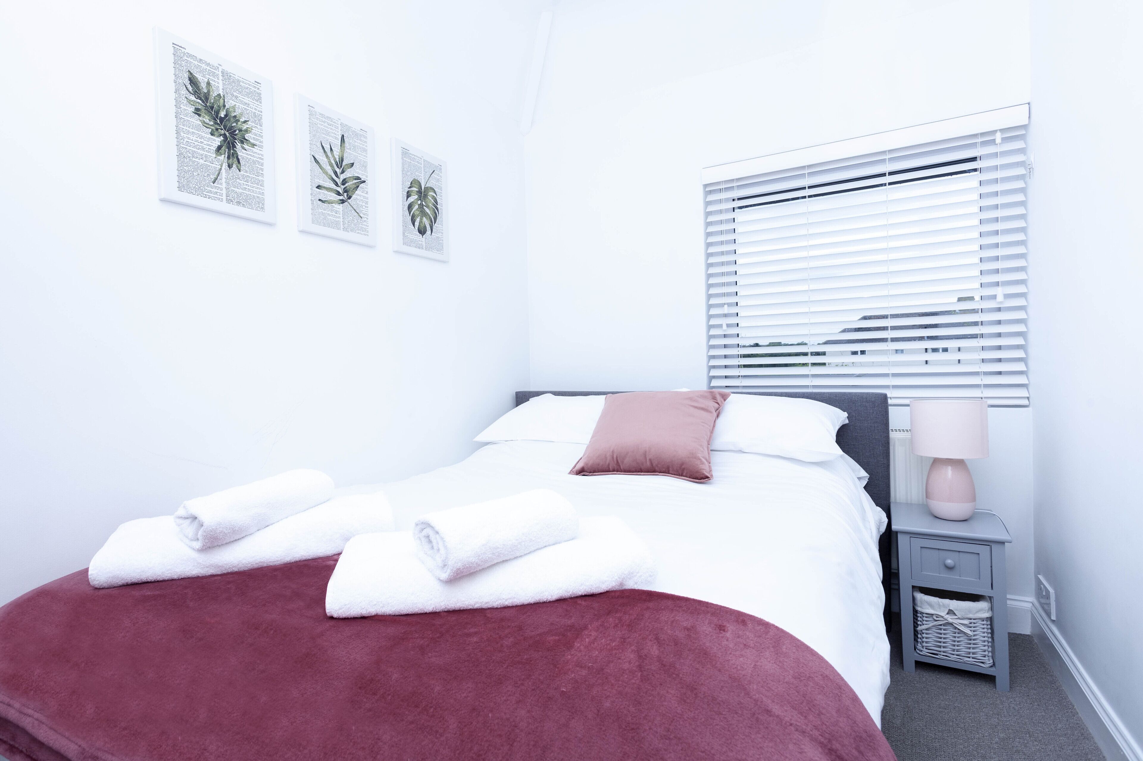 A neatly made bed with white and pink bedding, a red throw blanket, and two rolled white towels. There are three framed botanical prints on the wall, and a bedside table with a lamp and a basket.