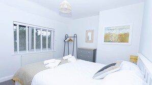 A neatly arranged bedroom with a bed, bedside tables, a dresser, a radiator, a window with blinds, and a hanging light fixture. - Magnolia Cove with 2 parking spaces (Whitstable)
