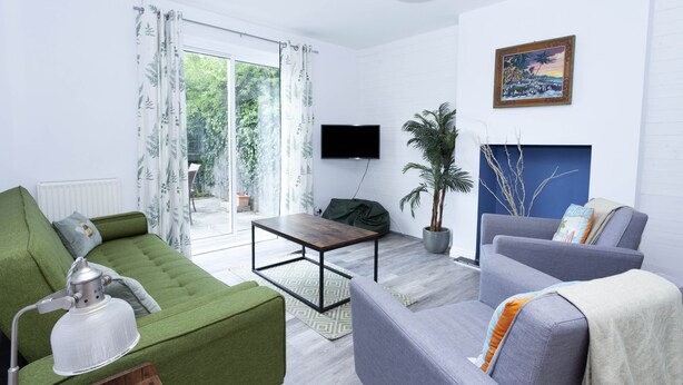 A modern living room with a green sofa, grey armchair, and a small table. There is a flat-screen TV mounted on the wall, a potted plant, and a fireplace with a blue accent wall. - Magnolia Cove with 2 parking spaces (Whitstable)