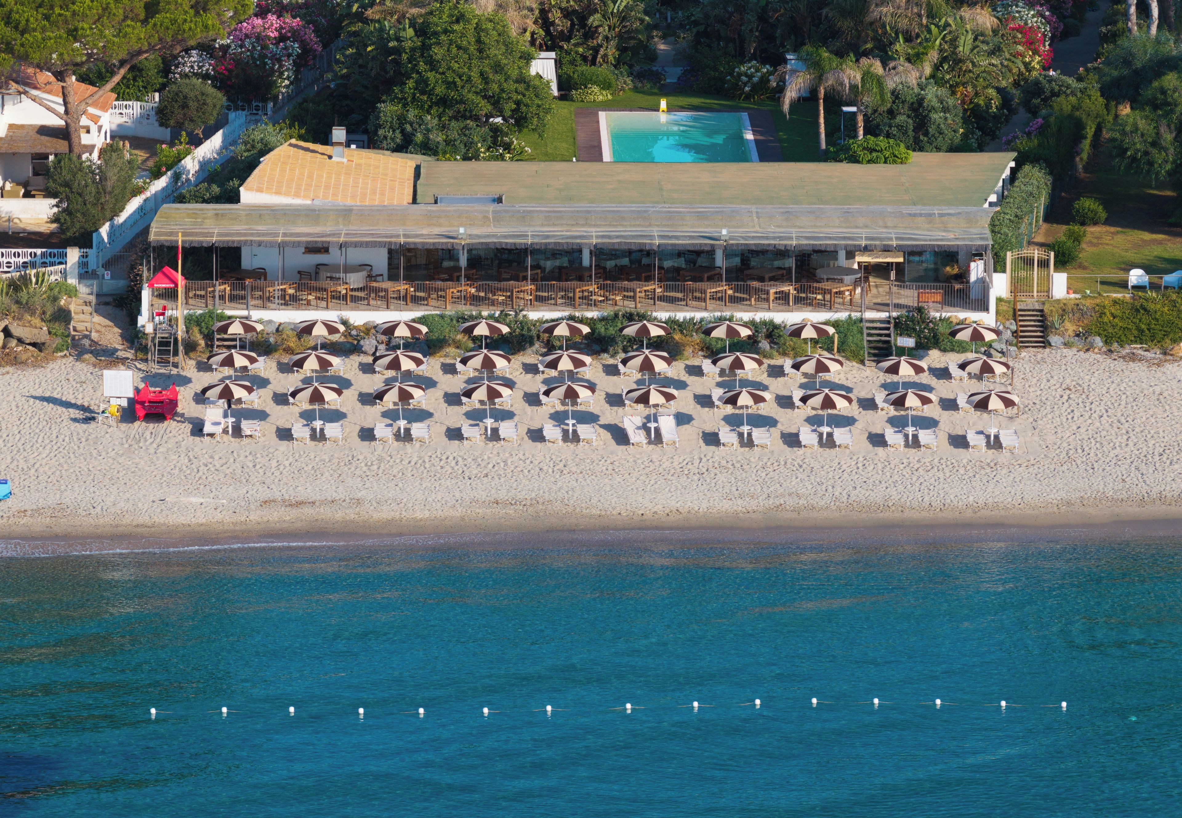 Vista aérea de un resort frente a la playa, con piscina, varias zonas de comedor y numerosas sombrillas y sillas de playa.