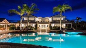 Una casa grande con piscina, palmeras y un patio bien iluminado.