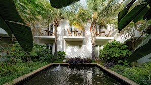 Un patio con una fuente central, rodeado de abundante vegetación y un edificio blanco con balcones.