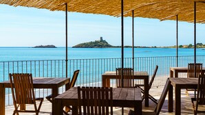 Un área de estar al aire libre con mesas y sillas de madera, techo de paja y vista al mar y una isla distante.