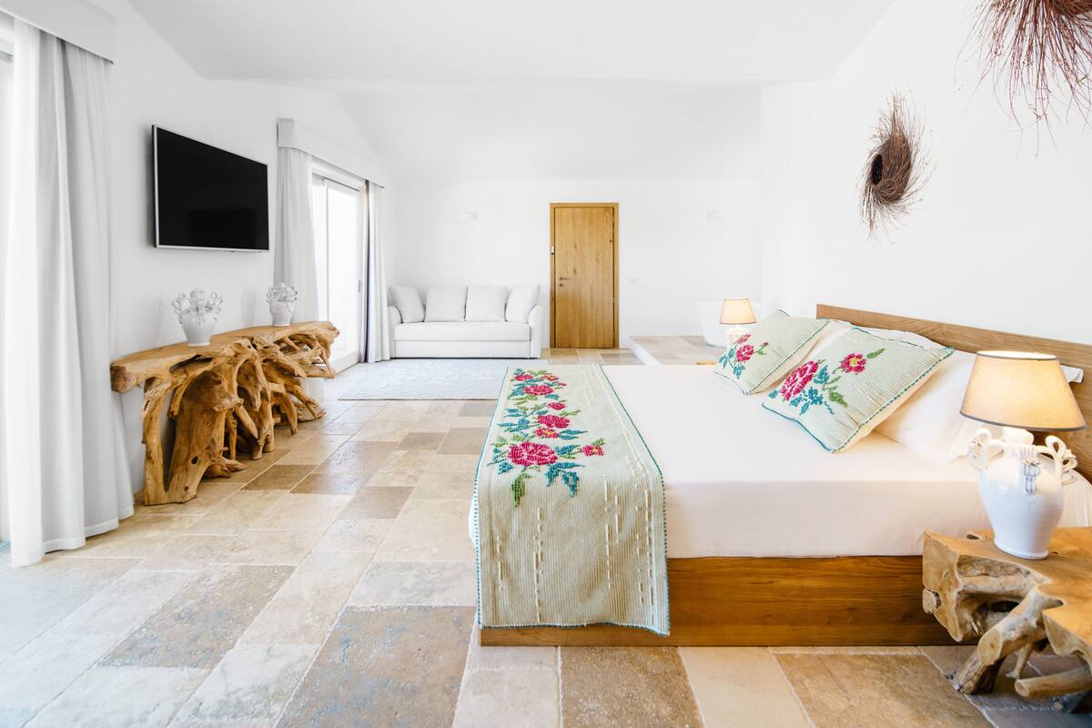 A bedroom with a bed, a wooden headboard, a bedside table with a lamp, a TV mounted on the wall, and a wooden bench with a cushion.