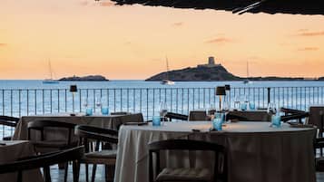 An outdoor dining area with tables set for two, overlooking a body of water with boats and a distant island.