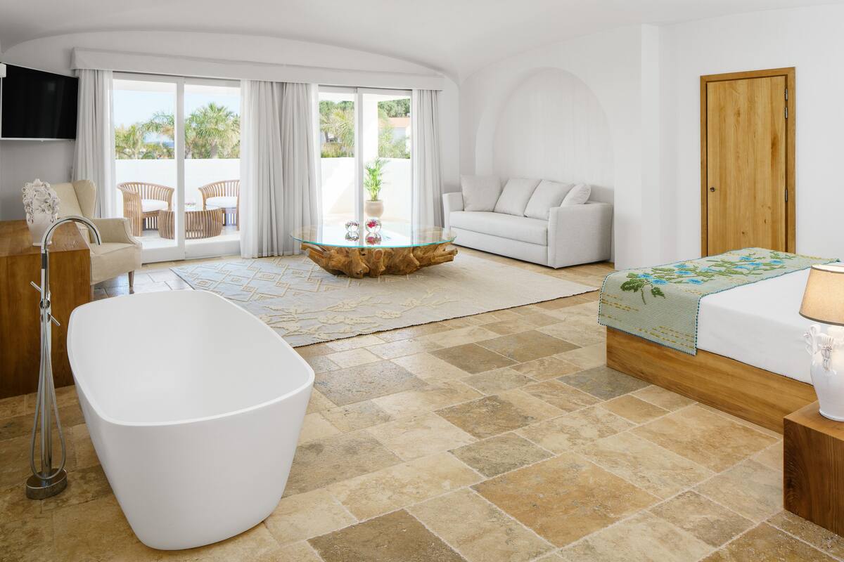 A modern hotel room with a large bathtub, a sofa, a dining table, and a wooden door.