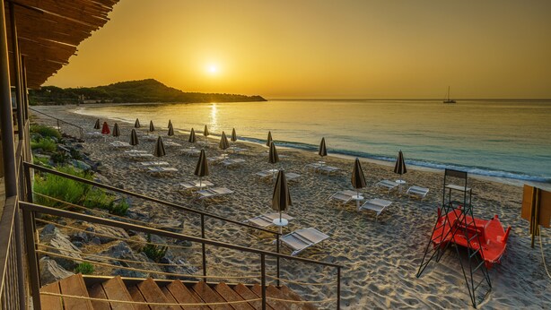 Una playa con tumbonas y sombrillas, una escalera de madera y vistas al océano al atardecer.
