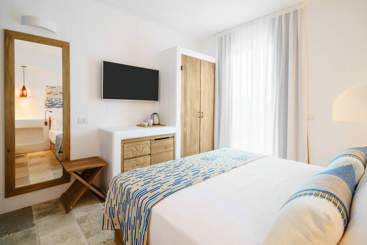 A hotel room with a large bed, a wooden wardrobe, a TV mounted on the wall, a small table with a kettle, and a mirror.