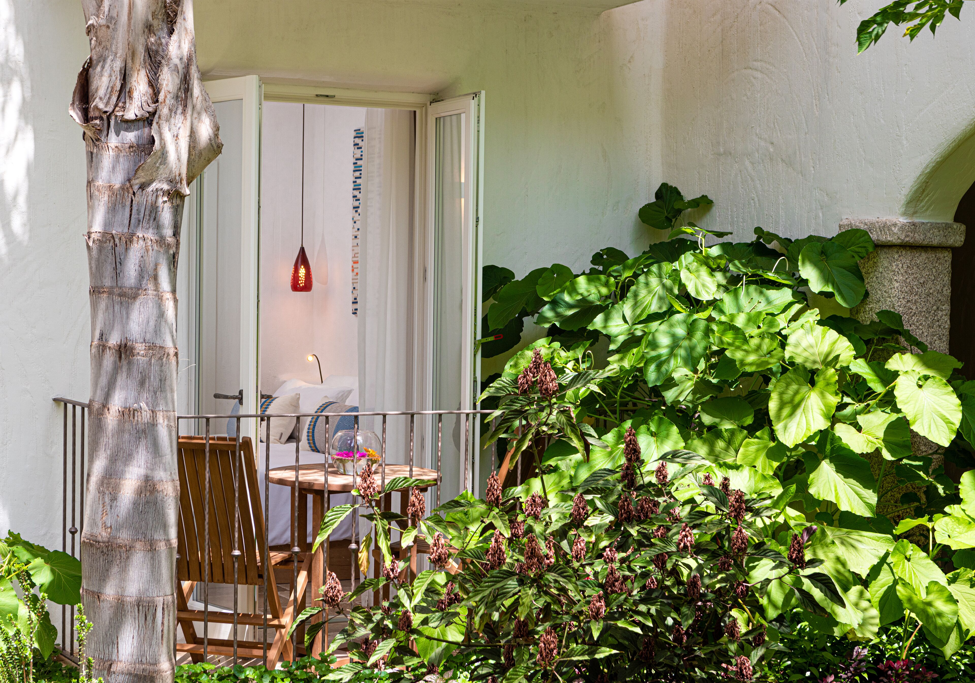 A patio with a wooden chair and table, a door with a hanging light, and lush green foliage.
