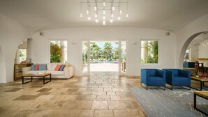 Un salón amplio con sofá, sillones azules y una mesa de centro. Un ventanal grande ofrece vistas a una piscina y palmeras.