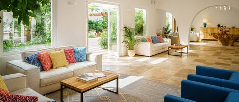 A spacious living room with a sofa, armchairs, and a coffee table. There is a large window providing a view of the outdoors.