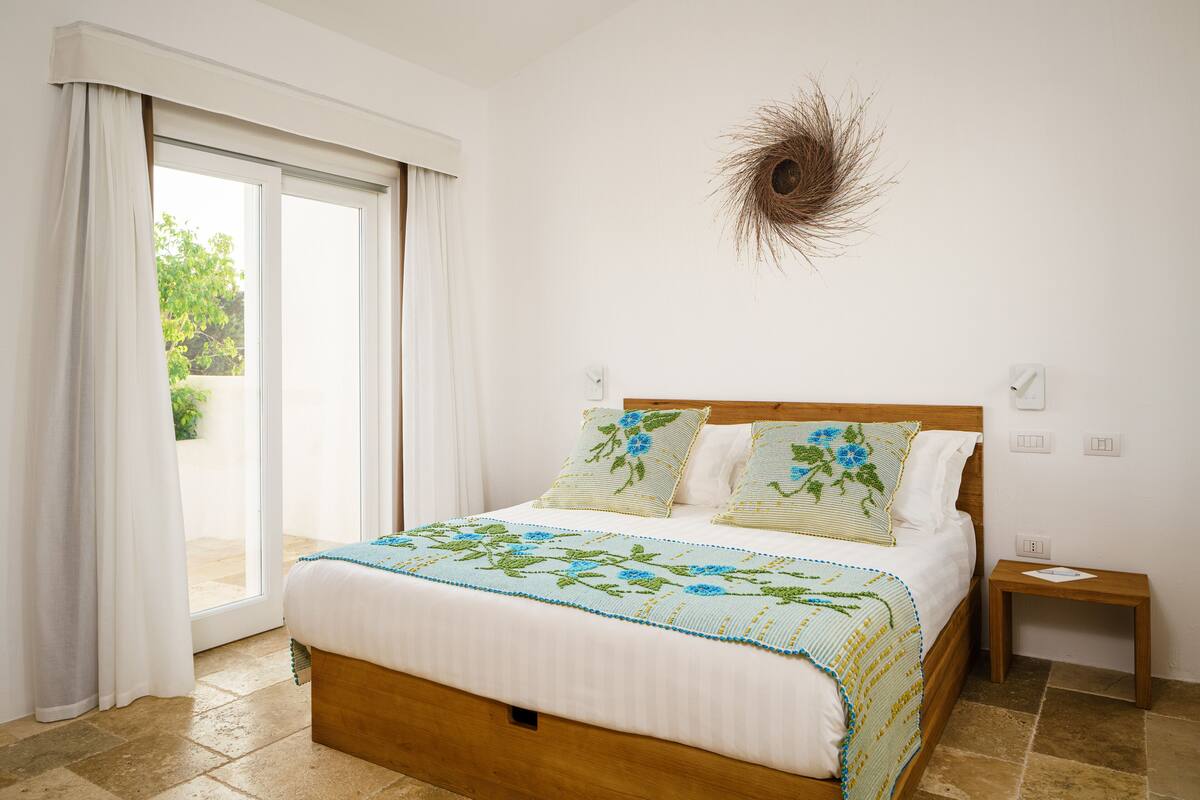 A bedroom with a bed, a nightstand, a wall-mounted light fixture, and a view through a sliding glass door.