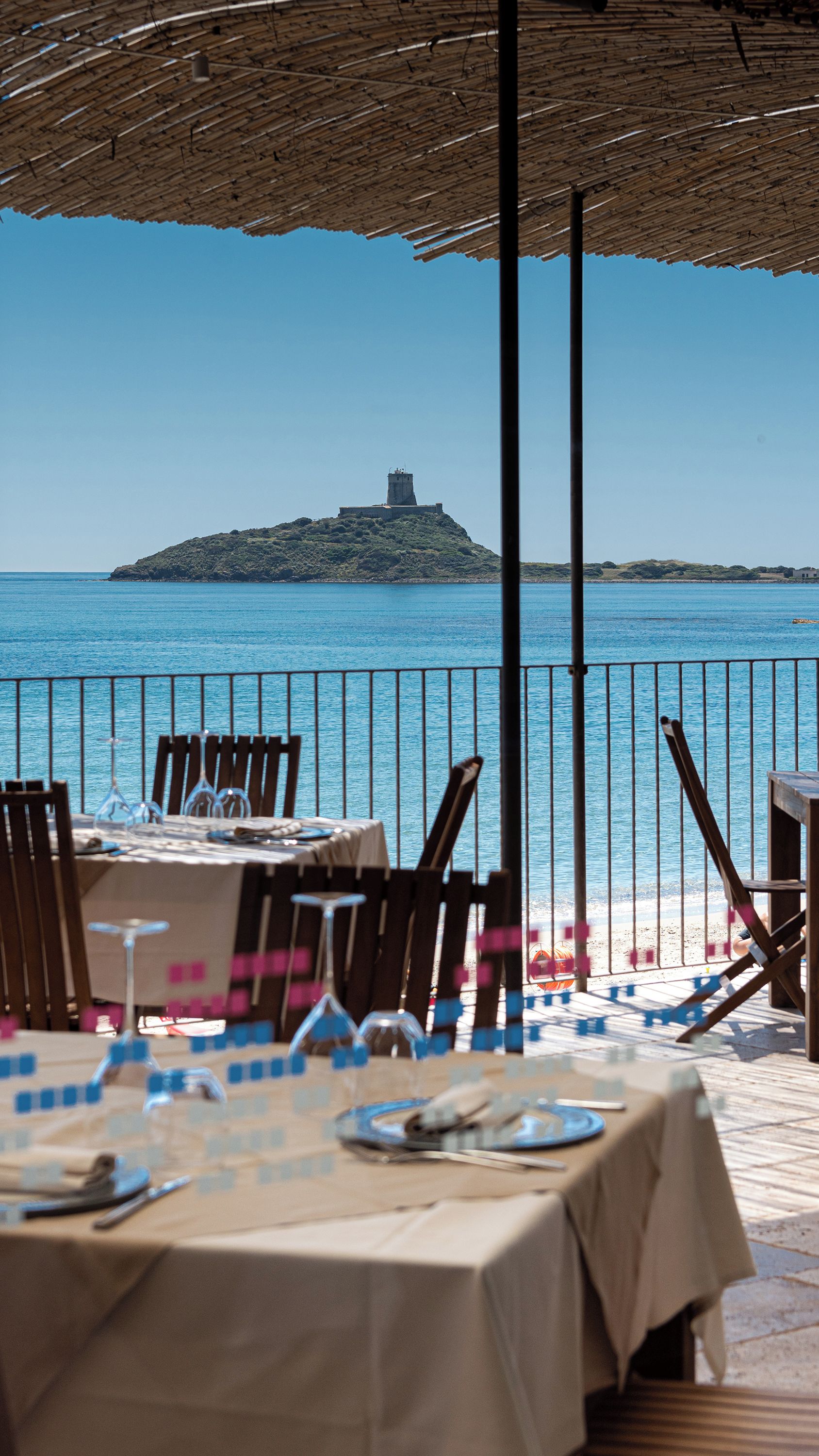 An outdoor dining area with tables set for two, overlooking a beach and a small island with a tower.
