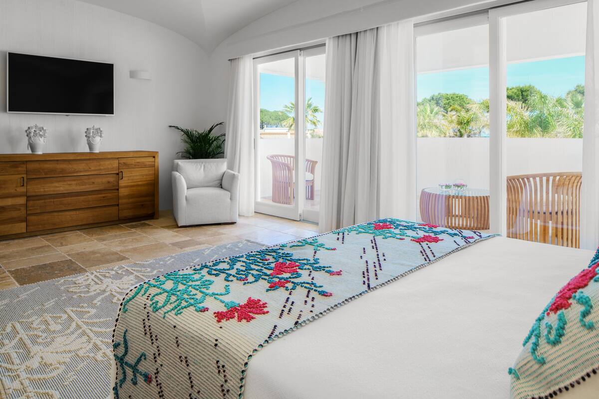 A hotel room with a large bed, a TV mounted on the wall, a wooden dresser, a potted plant, and a balcony with a view of the outdoors.