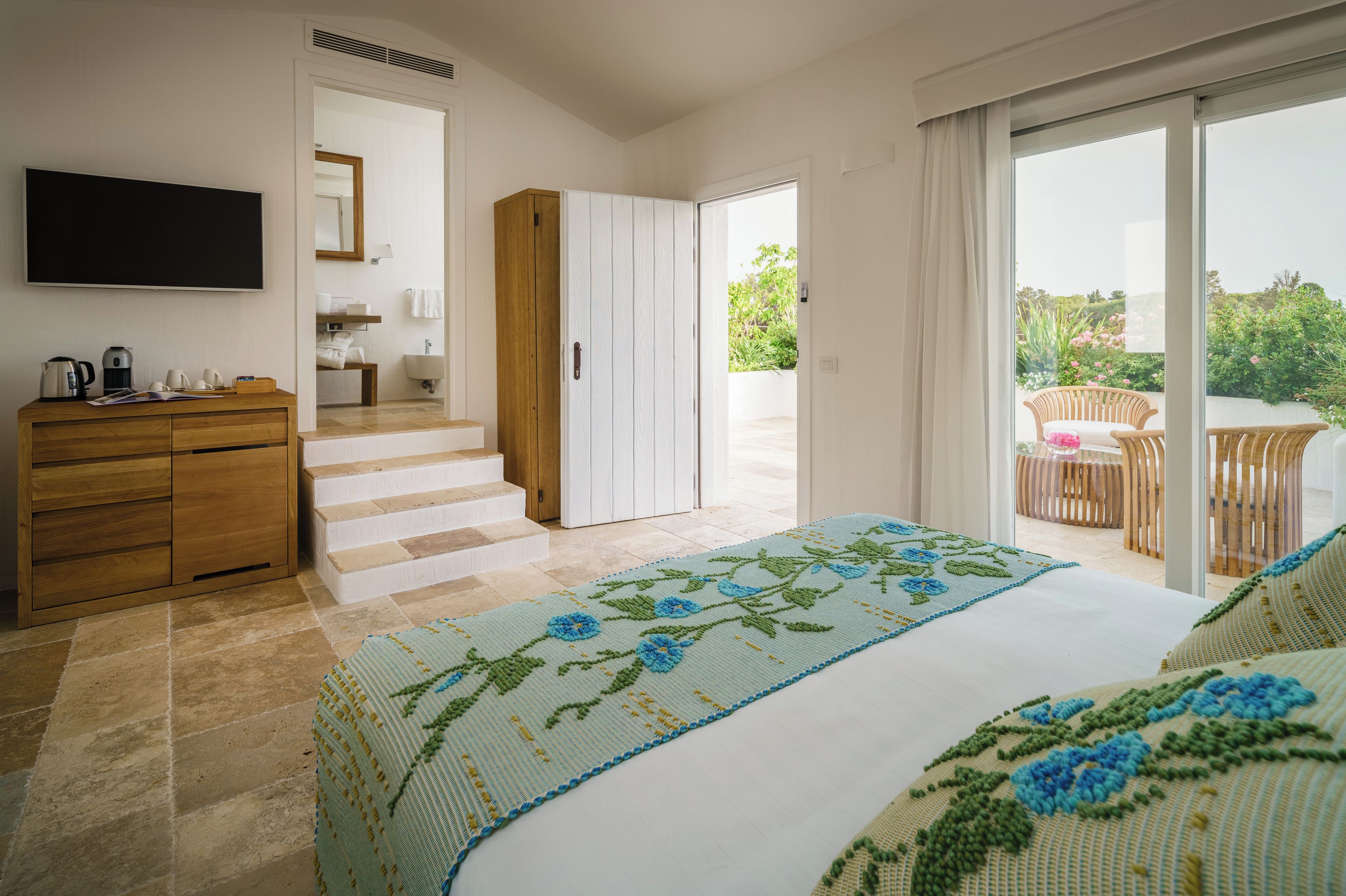 Une chambre à coucher avec un lit, une commode en bois, un téléviseur et une salle de bain visible à travers une porte ouverte.