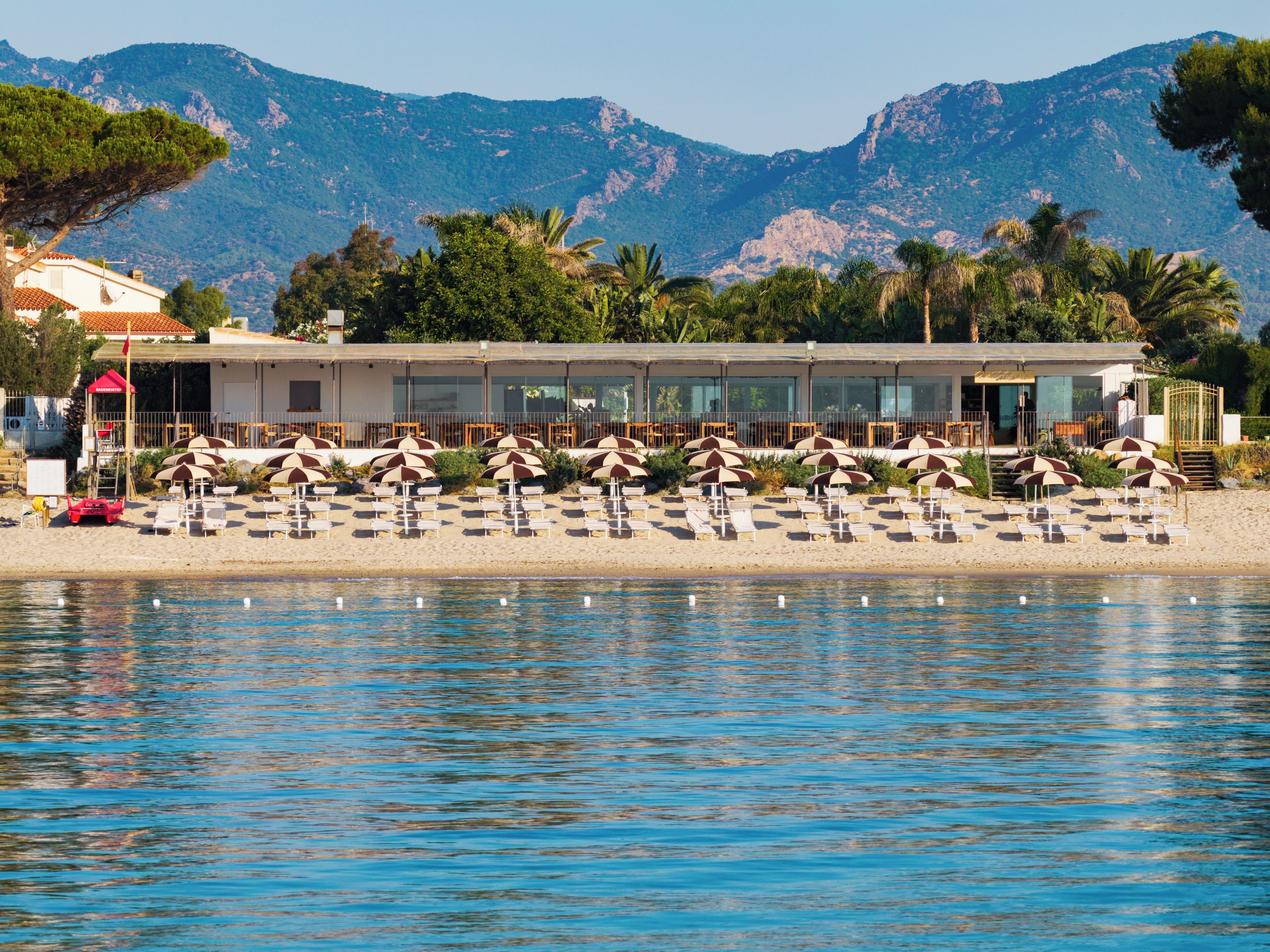 Ein Hotel direkt am Strand mit Liegestühlen, Sonnenschirmen, einem Pool und Bergen im Hintergrund.