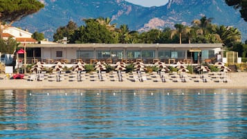 Ein Hotel direkt am Strand mit Liegestühlen, Sonnenschirmen, einem Pool und Bergen im Hintergrund.