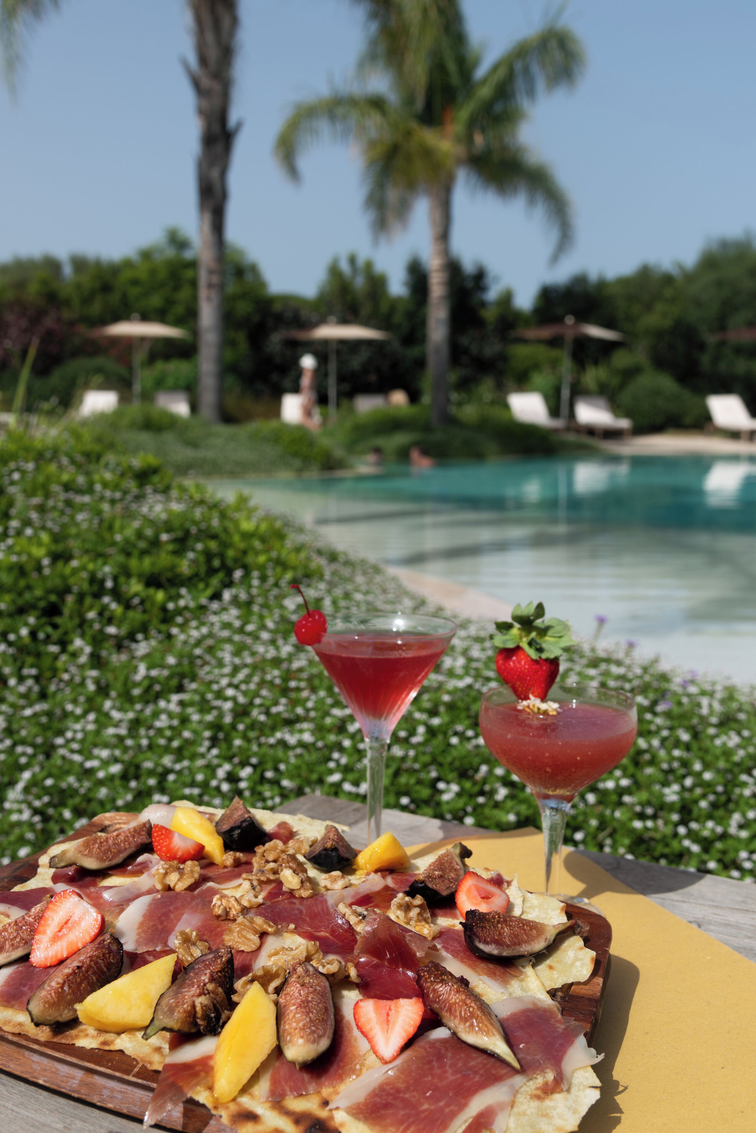 Ein Frühstücksteller mit Früchten, Nüssen und Käse, dazu zwei Cocktails auf einem Tisch am Pool.