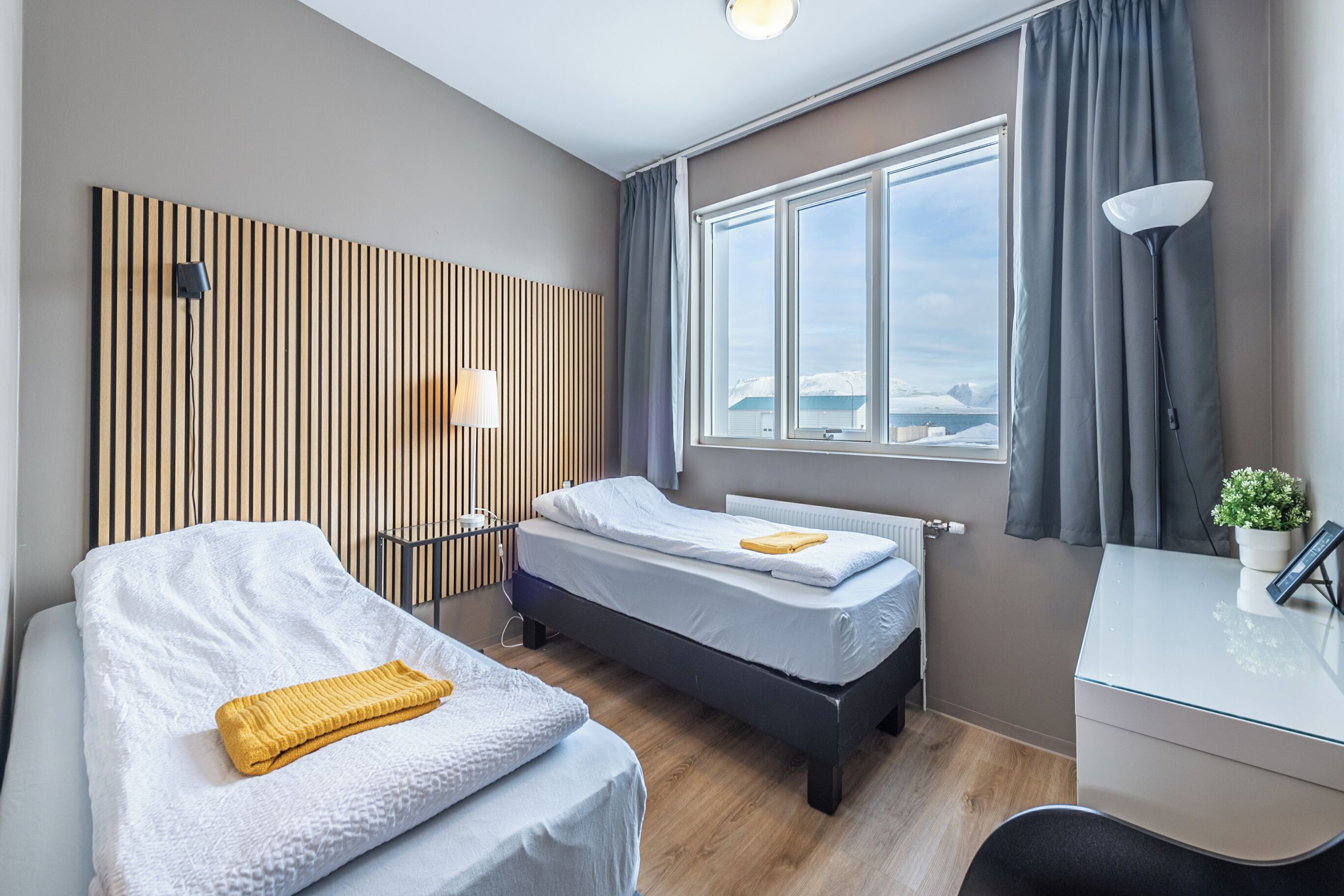 A modern hotel room with two beds, a desk, a lamp, and a view of the sea.