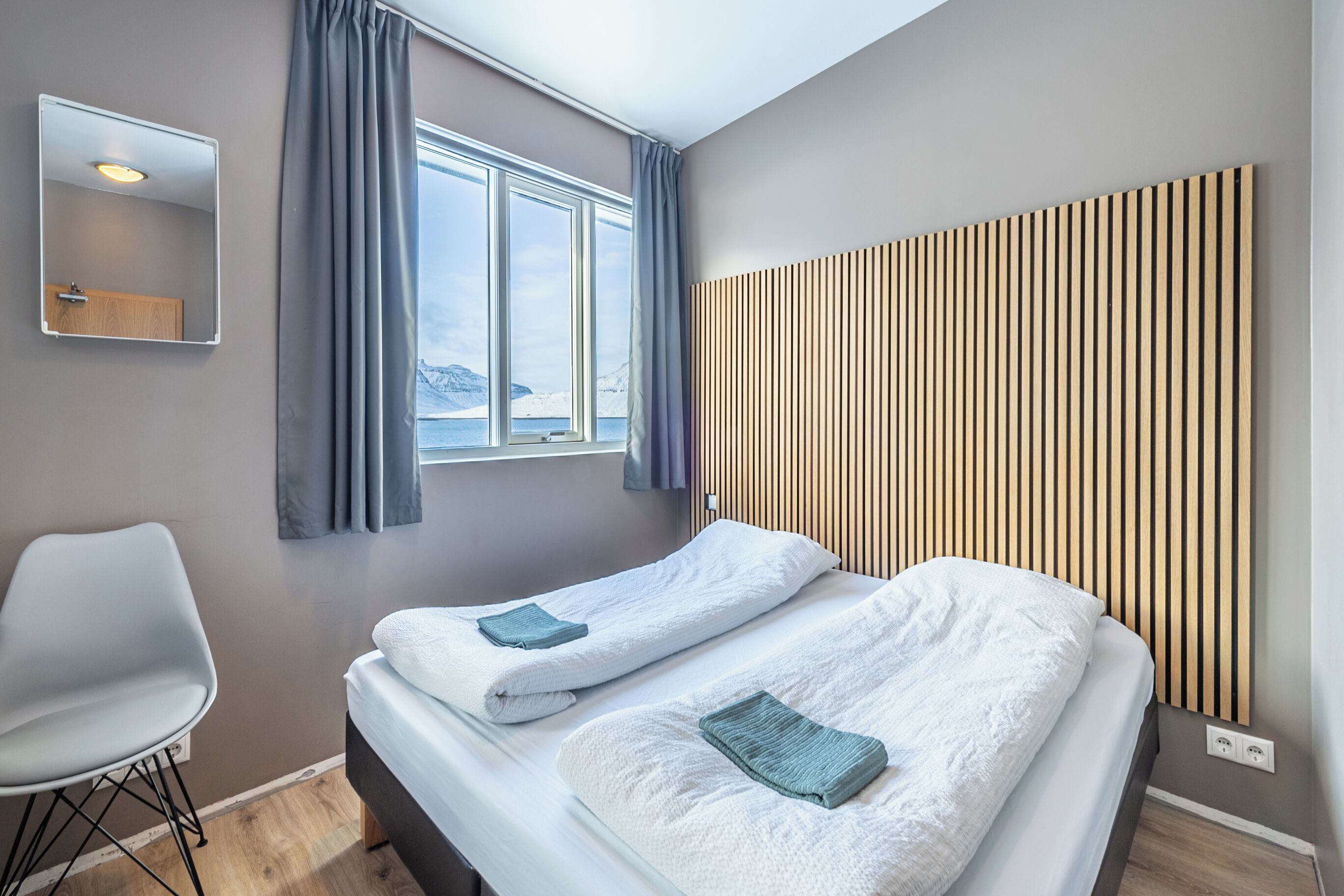 Quarto de hotel moderno com duas camas, uma janela grande, cabeceira de ripas de madeira, espelho e uma cadeira.