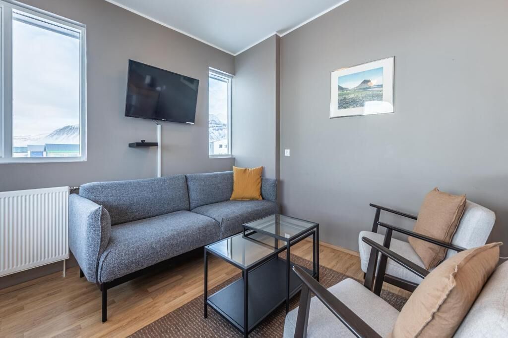 Un salon moderne avec un canapé gris, une table basse en verre et un fauteuil blanc. Un téléviseur à écran plat est fixé au mur, et un tableau encadré est visible.
