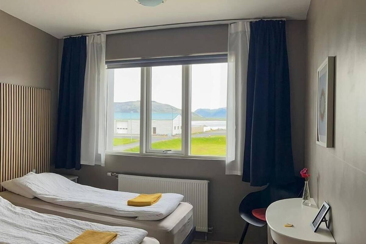 A hotel room with two beds, a small table, a chair, a window with a view of a building and landscape, and a radiator.