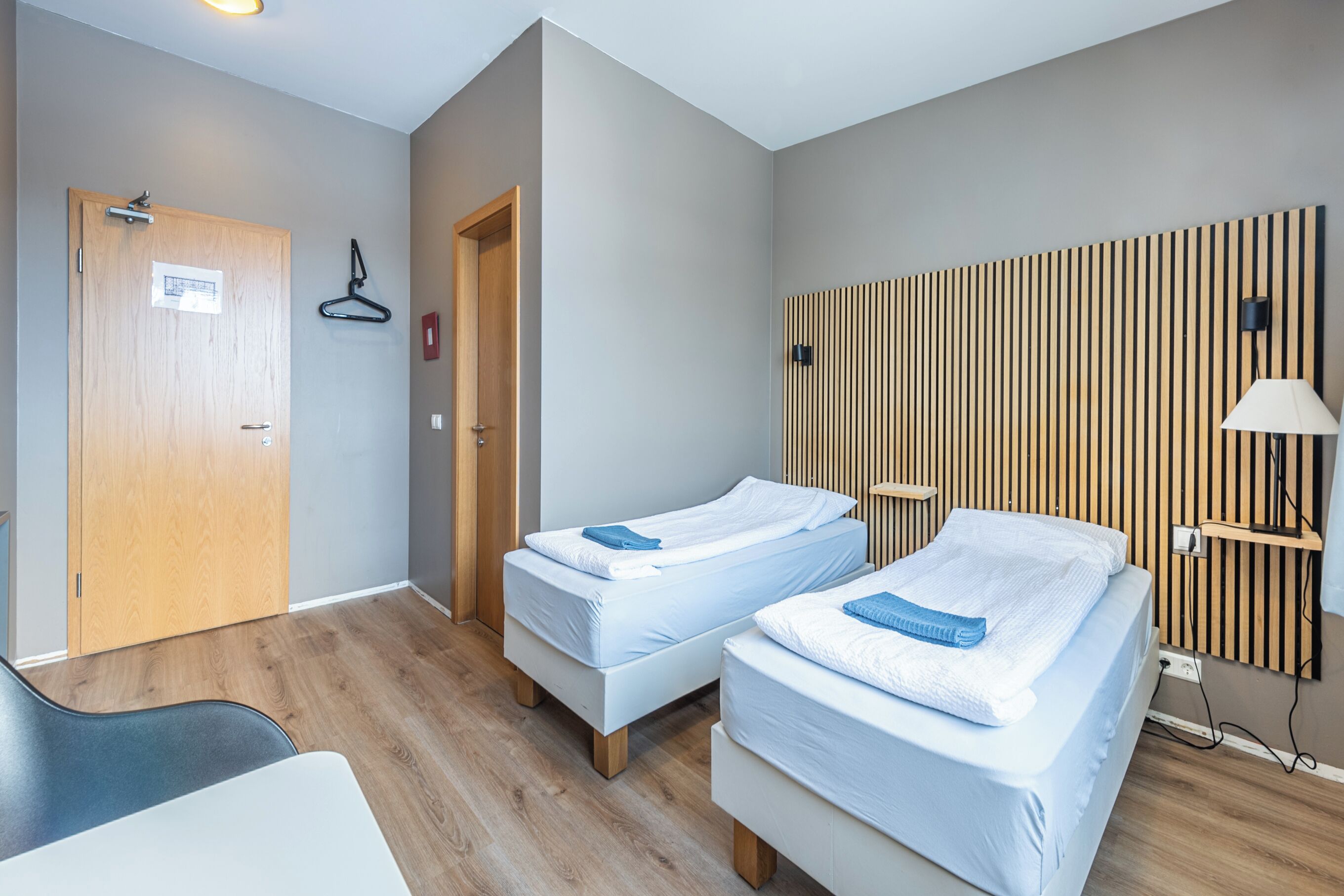 Habitación de hotel con dos camas individuales, una puerta de madera, un perchero y una lámpara fijada a la pared.