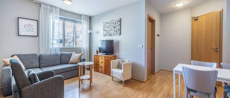 Een moderne woonkamer met een grijze bank, een klein rond tafeltje, een witte stoel en een televisie op een houten kast.