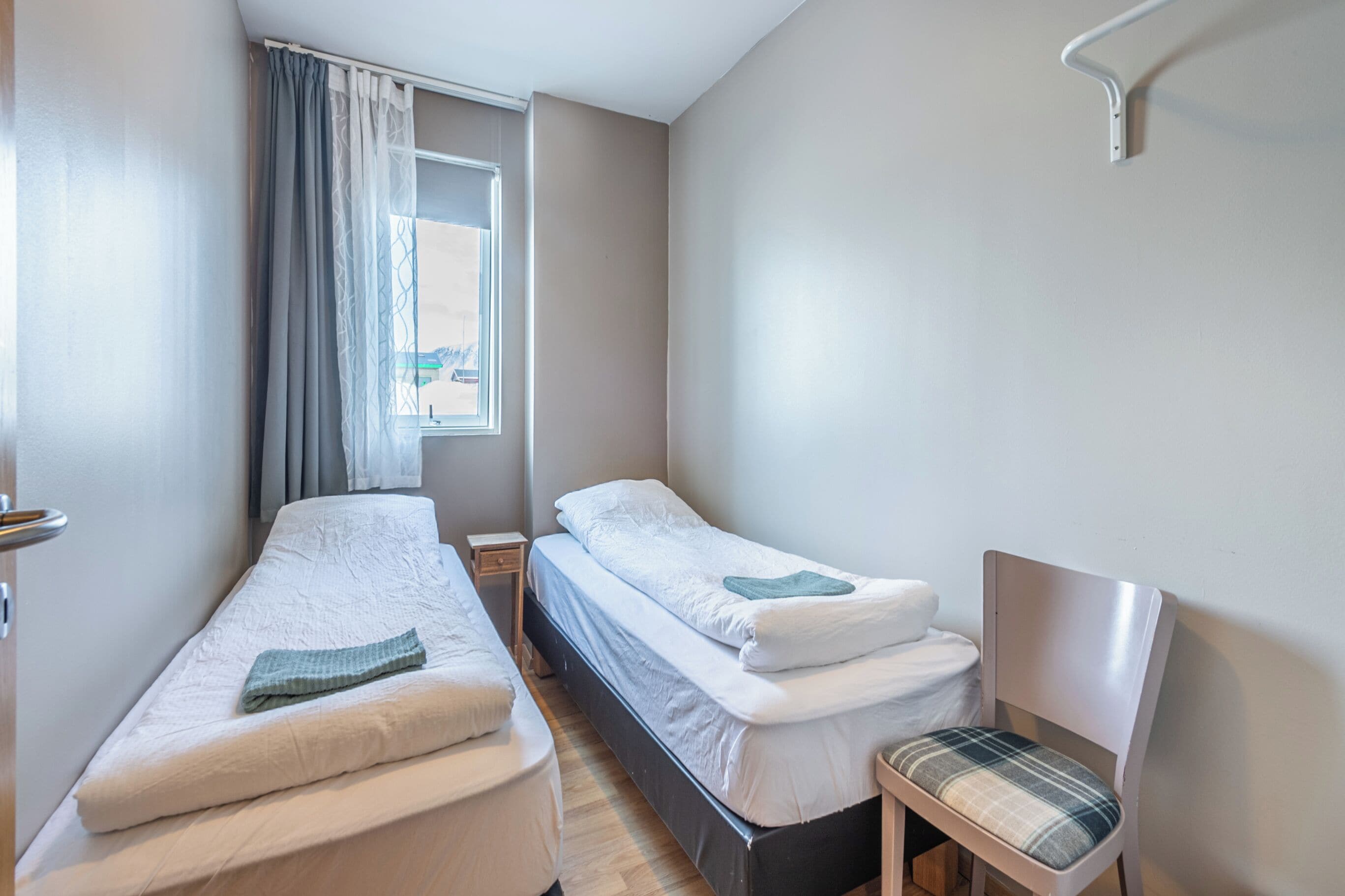 A small, clean room with two single beds, a chair, and a window with curtains.