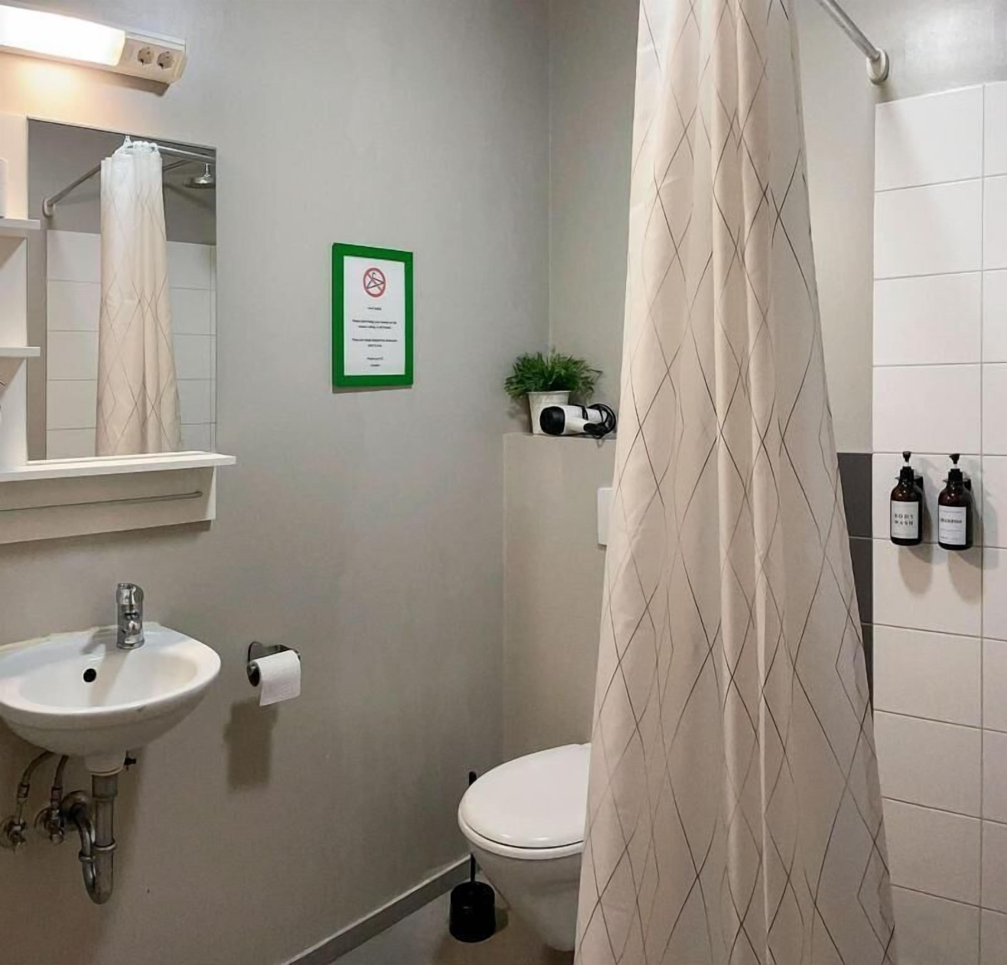 Un baño con lavamanos, inodoro y cortina de ducha. Hay un letrero verde en la pared y una planta en un estante.