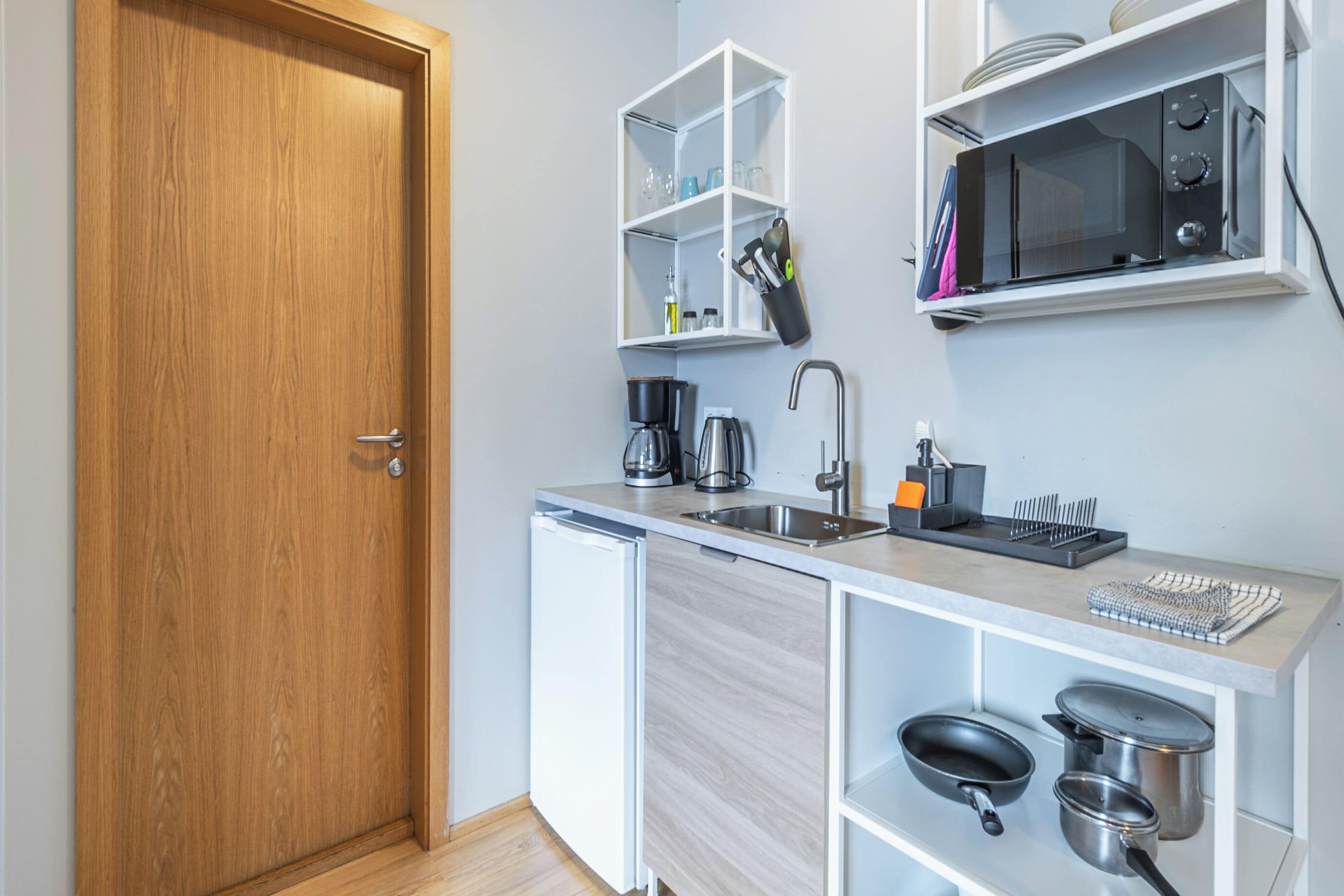 A compact kitchen with a wooden door, white cabinetry, a built-in microwave, a coffee maker, and a sink.