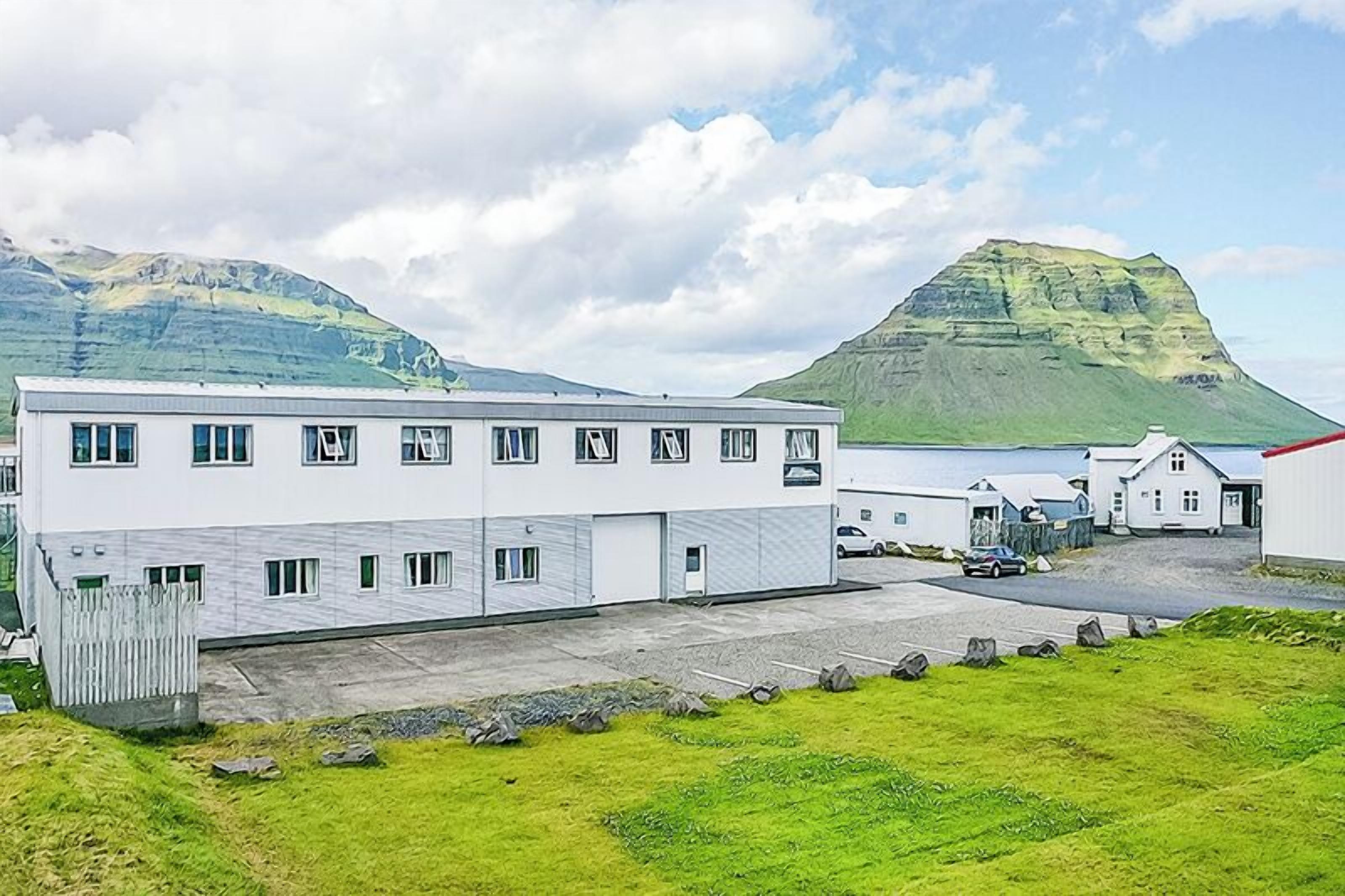 A modern building with multiple windows and a parking area, set against a backdrop of mountains and a partly cloudy sky.
