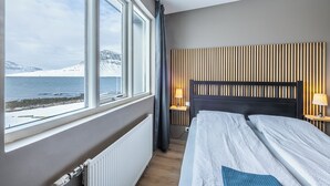 Habitación de hotel con un amplio ventanal que ofrece vistas a un paisaje nevado y una masa de agua. La habitación tiene una cama con cabecero de madera, dos mesitas de noche con lámparas y un radiador.