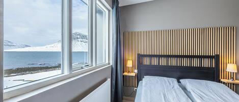 A hotel room with a large window offering a view of a snowy landscape and a body of water. The room features a bed with a wooden headboard, two bedside tables with lamps, and a radiator.
