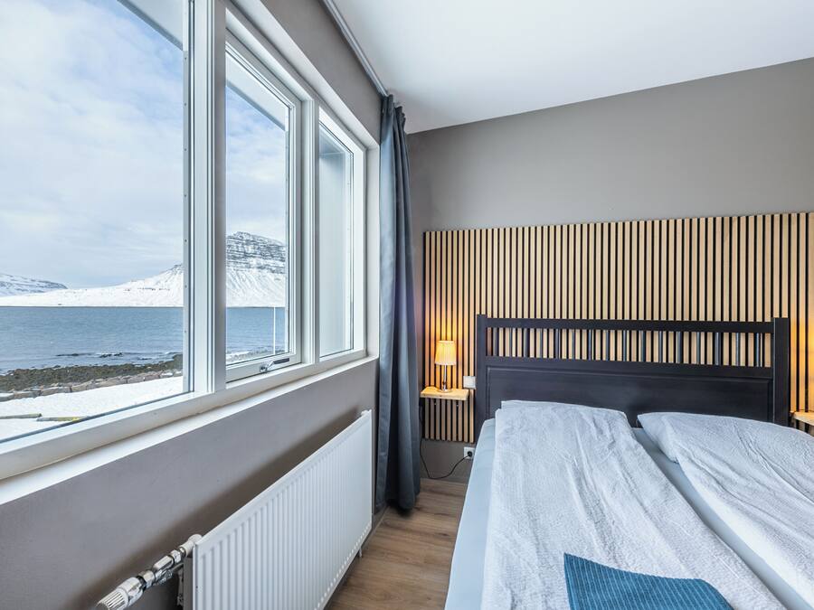 A hotel room with a large window offering a view of a snowy landscape and a body of water. The room features a bed with a wooden headboard, two bedside tables with lamps, and a radiator.