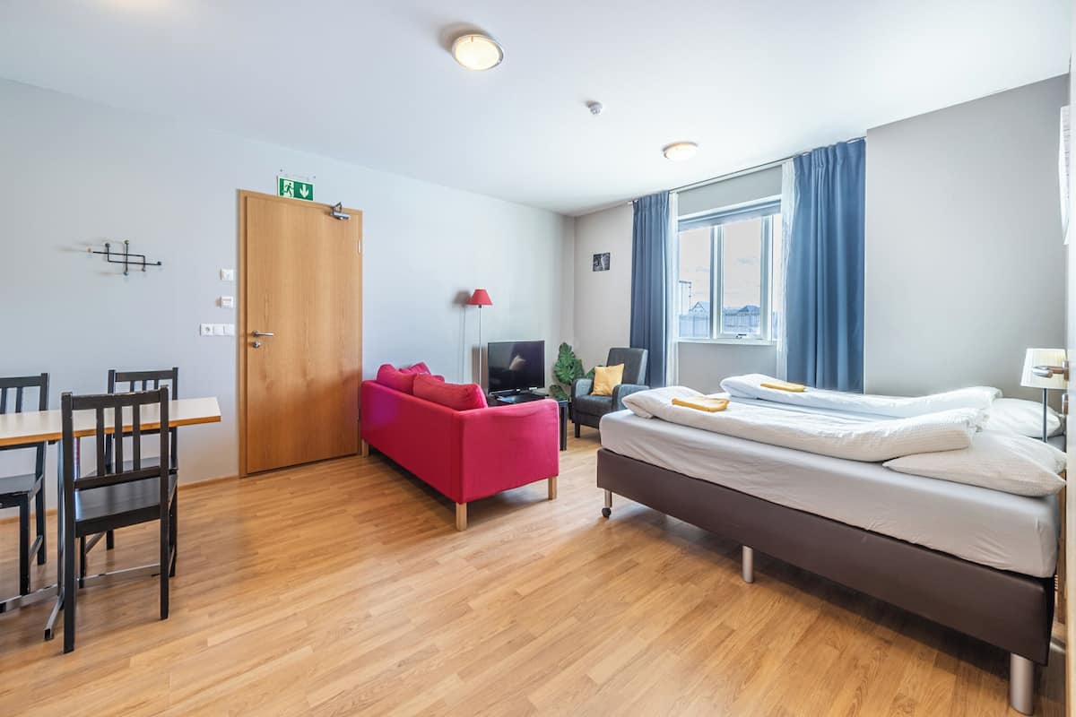 A modern hotel room with a wooden floor, a bed, a red sofa, a TV, and a window with curtains.
